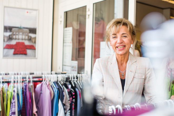 Foto: Eine Mitarbeiterin des Rotkreuz-Shops steht zwischen mehreren Ständern mit farbenfroher Kleidung. Sie blickt lachend in die Kamera.
